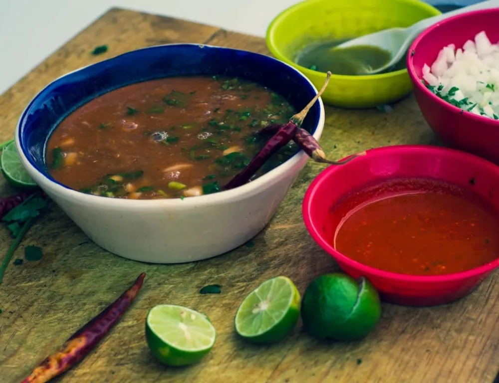 Consomé de birria con cilantro, chile seco, salsa roja, cebolla y limones