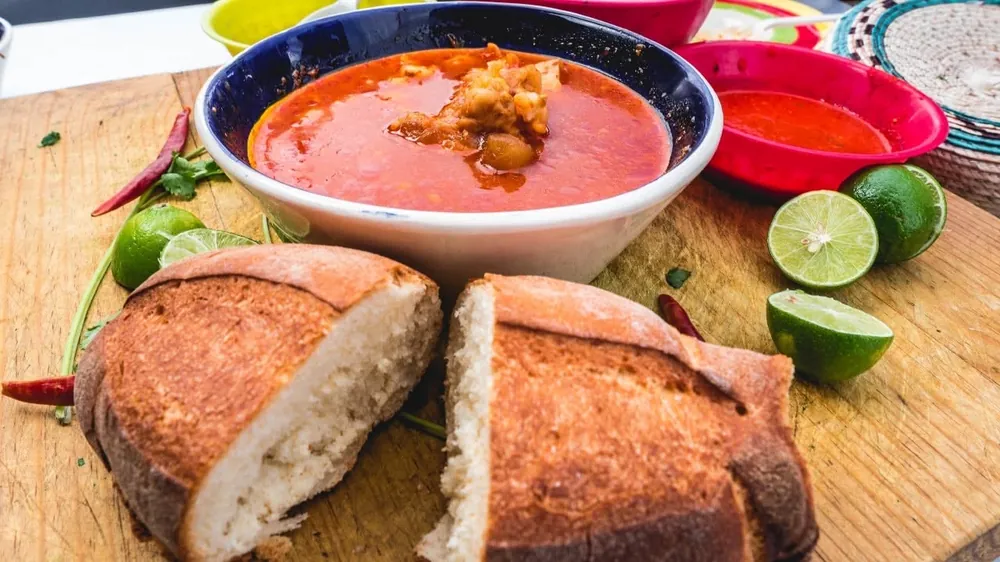 Plato de menudo rojo con pan, limones y chile de árbol